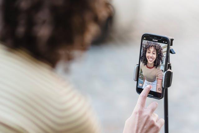 A young woman presses the camera record button on her phone that’s on a tripod. 
