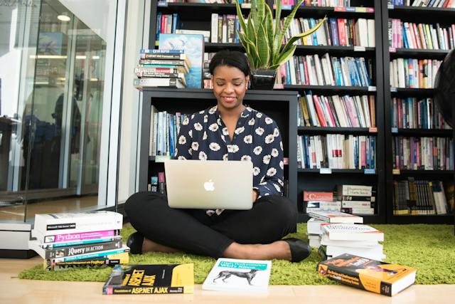 Une femme est assise par terre, utilisant son ordinateur portable et entourée de livres.