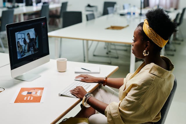 A woman sits on a desk and browses through video clips on a computer. 
