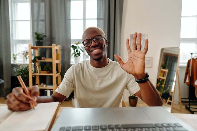 A man sits in front of a laptop and waves his hand to greet his viewers. 