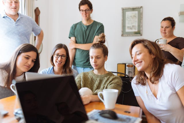 O imagine a unui grup de persoane care stau în picioare și în picioare în jurul unei mese și urmăresc videoclipuri TikTok pe un laptop. 
