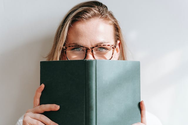A woman wearing glasses peeks over a book. 