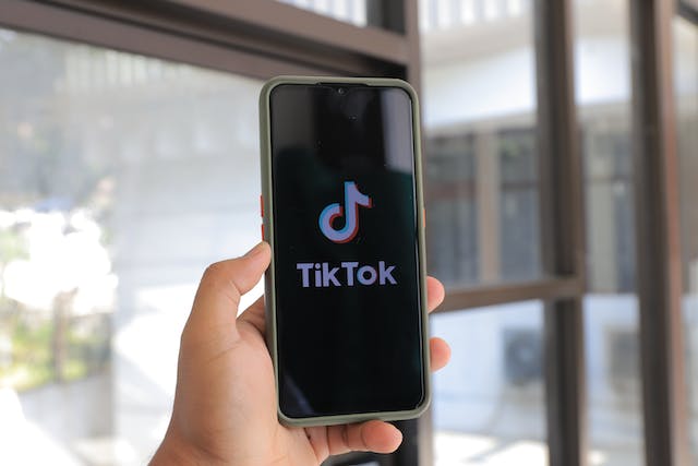 A person holds up their phone with the screen displaying the TikTok logo and name. 
