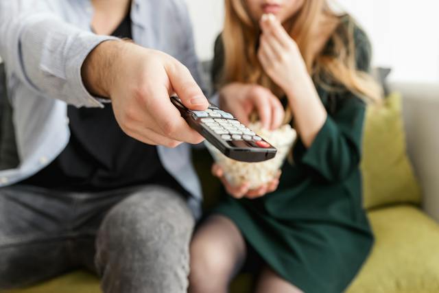 A man uses a TV remote control while sharing a bowl of popcorn with a woman. 