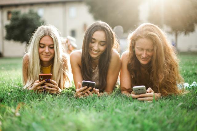 Three girls lie on the grass while browsing on their phones. 