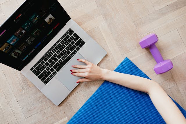 Een vrouw op een gymnastiekmat zit op haar laptop en heeft een gewicht naast zich.