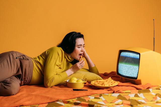  Imagem de uma mulher deitada em frente a uma TV enquanto come um lanche. 