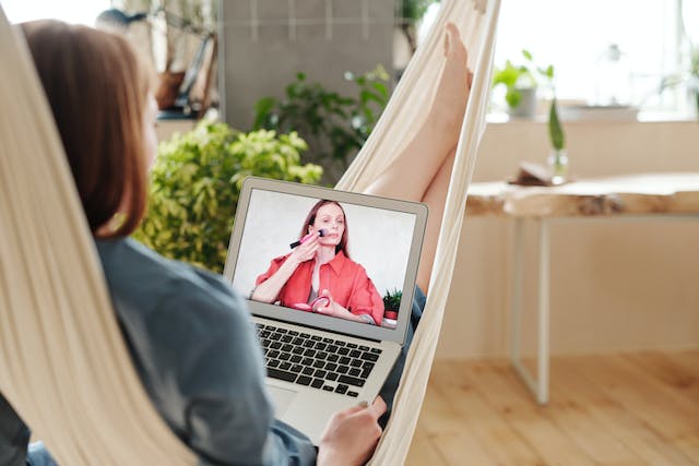 A woman lies on a hammock and watches a TikTok makeup tutorial on her laptop. 
