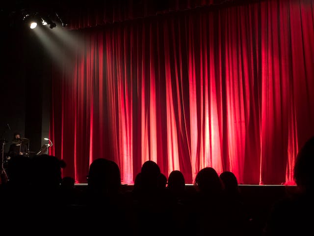 A theater audience waits for the red curtains on the theatre stage to open. 
