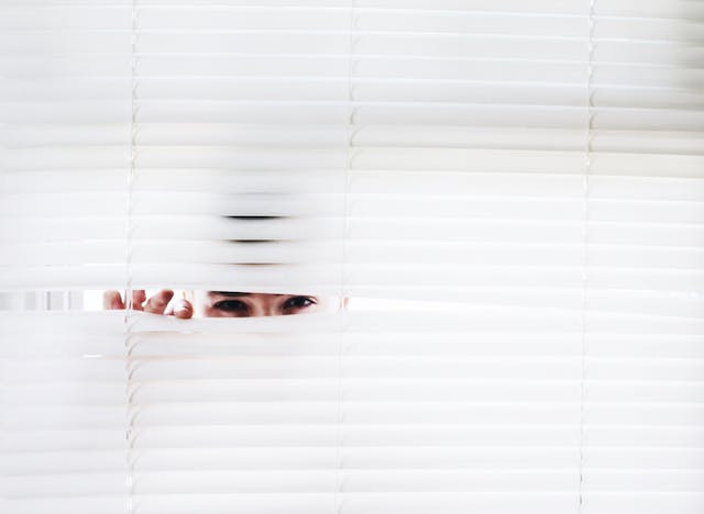 Uma pessoa está espiando através das persianas brancas da janela. 