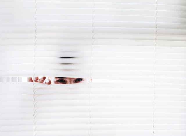 Une personne regarde à travers les stores blancs d'une fenêtre. 