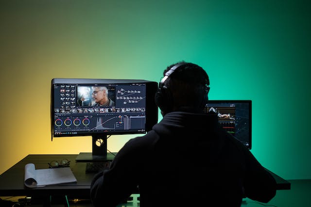 A man wearing headphones sits in front of two monitors and edits videos on his computer. 
