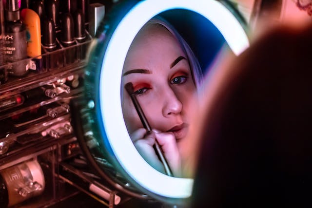 Een vrouw zit voor een spiegel en gebruikt een kwast om oogschaduw aan te brengen. 