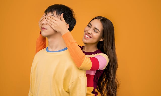 Een vrouw staat achter een man en bedekt zijn ogen met haar handen. 