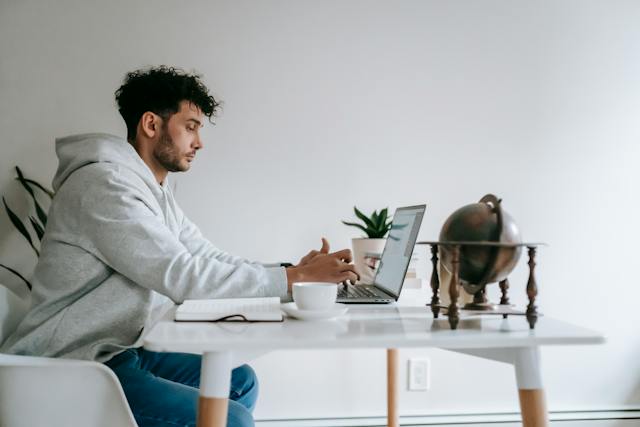 Um homem está sentado em uma mesa enquanto trabalha em seu laptop. 