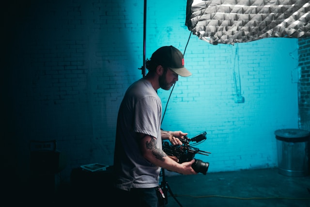 A man in a white T-shirt and a cap holding a black camera.
