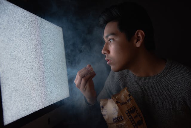 A man eats popcorn as he stares at static on a big TV monitor. 
