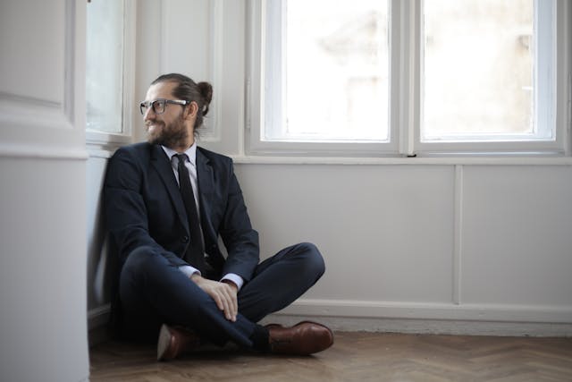 A man wearing glasses and a suit sits on the floor while he peeks out a window. 
