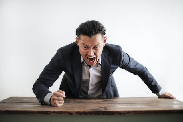 Un homme se penche sur une table, le poing levé, et crie à la caméra.