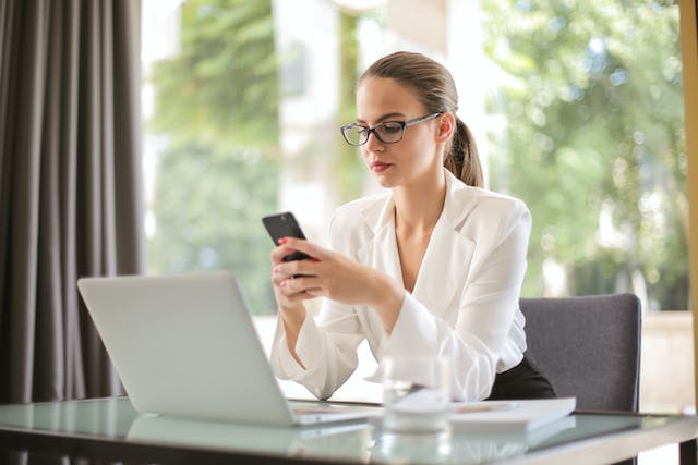 Een vrouwelijke professional zit aan een kantoortafel terwijl ze op haar telefoon door de commentaren van TikTok bladert. 