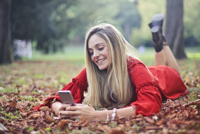 Een vrouw ligt op haar buik op gevallen bladeren terwijl ze op haar telefoon scrollt.
