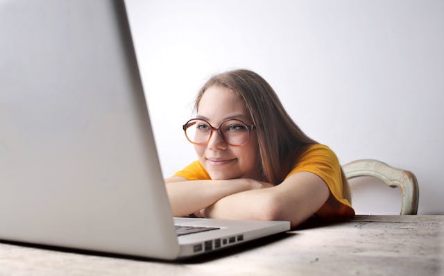 A smiling woman rests her arms and head on a table while staring at her laptop. 
