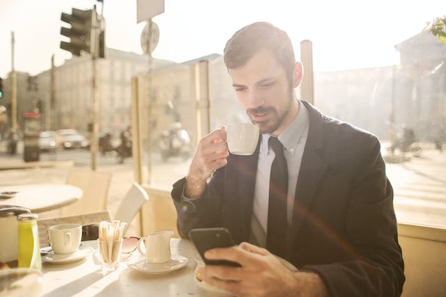 A man drinks coffee at an outdoor cafe while he scrolls on his phone. 
