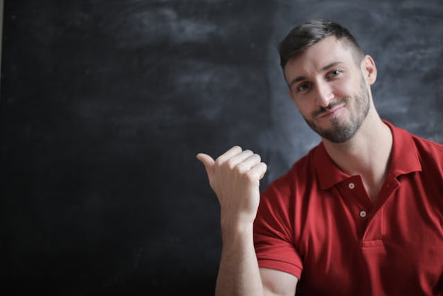 A man in a red shirt gestures with his thumb as a sign of invitation. 
