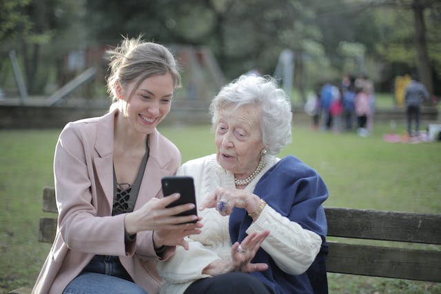 A woman shows videos on her phone to her grandma while they’re sitting on a park bench. 
