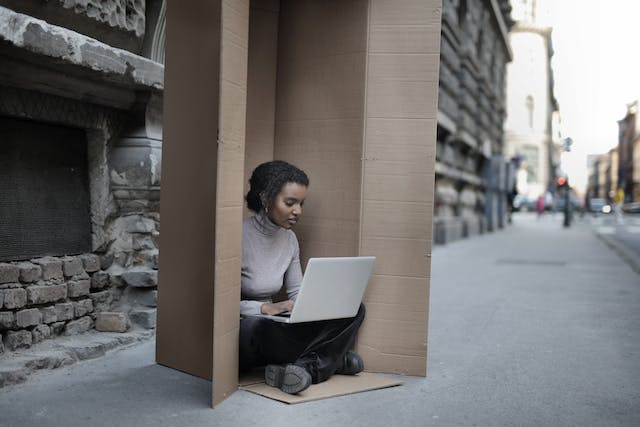 A woman sits inside a large cardboard box while using her laptop. 