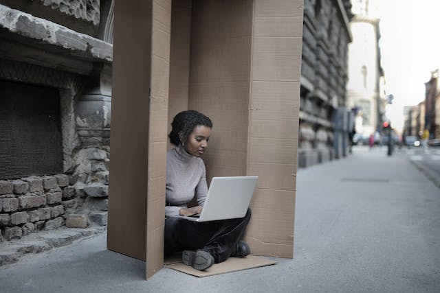 O femeie stă în interiorul unei cutii mari de carton în timp ce își folosește laptopul. 