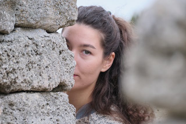 A woman hides half her face behind a wall of big stone blocks. 
