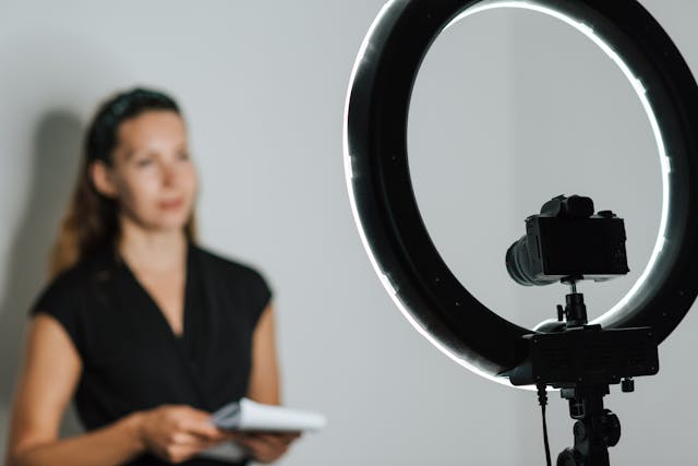 A woman holds a notepad and stands in front of a ring light and a digital camera. 
