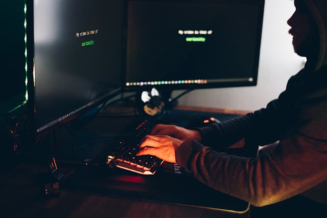 Un bărbat stă în întuneric în fața a două monitoare de calculator și tastează la tastatură. 