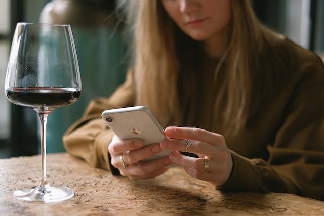 A woman browses TikTok on her phone as she drinks a glass of red wine. 
