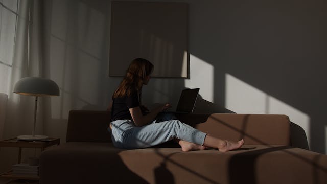 Una donna siede sul divano nell'ombra e sfoglia i video di TikTok sul suo portatile. 