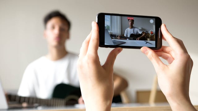 Iemand die een telefoon gebruikt om op te nemen dat iemand anders gitaar speelt.