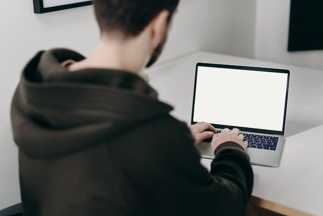 A man types on his laptop, which displays a blank screen. 
