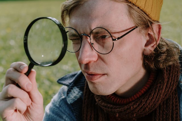 Um homem de óculos segura uma lente de aumento em um dos olhos. 