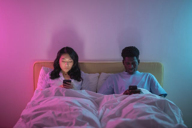 A couple lies side by side in bed as they scroll through their phones. 
