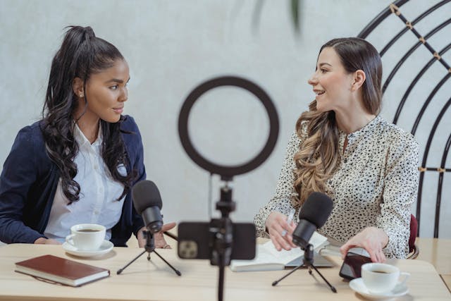 Duas mulheres sentam-se em frente a microfones em uma mesa e a uma câmera de telefone e participam de uma discussão animada durante uma transmissão ao vivo.