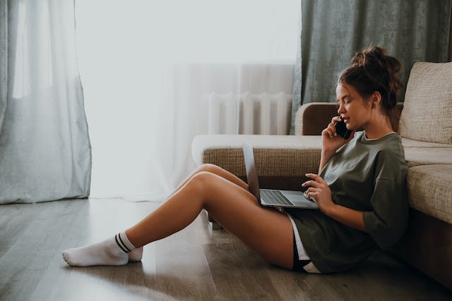 女性が床に座り、電話で話しながらノートパソコンを見ている。