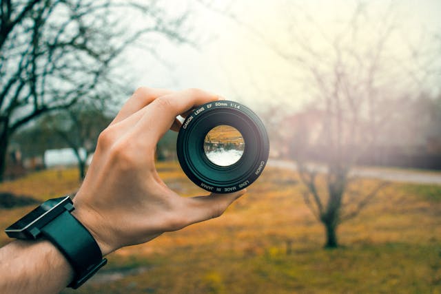 A person holds a DSLR camera lens over a view of a lake. 
