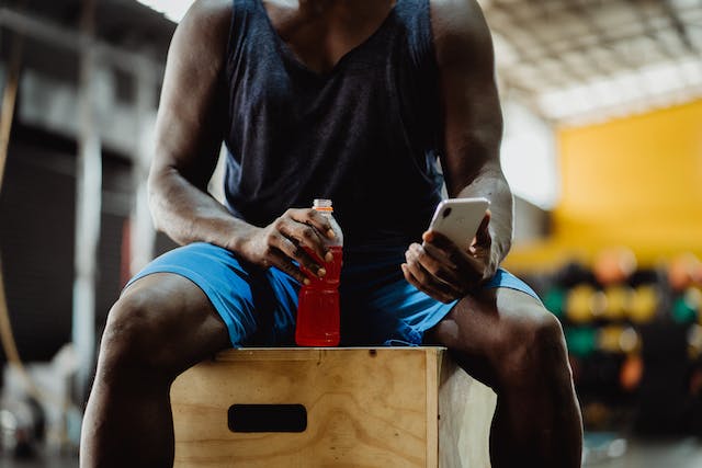 A man holds an energy drink, sits on a crate, and browses online on his phone. 
