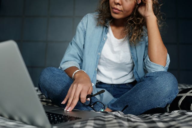 A woman sits on the bed looking frustrated while browsing on her laptop. 
