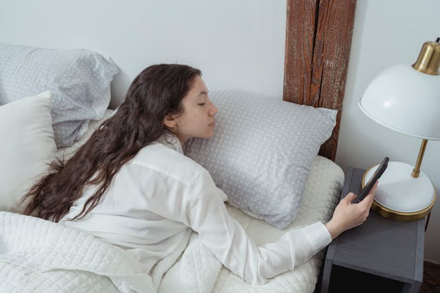 A woman lays in bed after she wakes up and checks her phone. 
