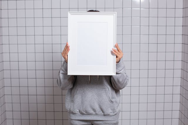 A person stands in a white, tiled room and hides behind an empty, white picture frame. 
