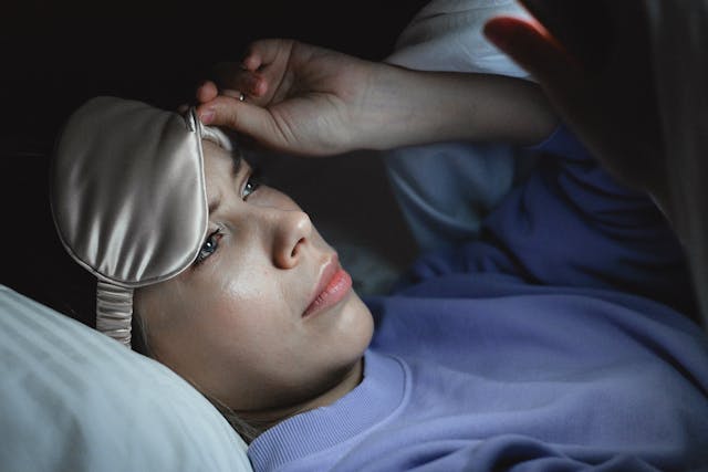 Eine Frau im Bett lugt unter ihrer Schlafmaske hervor und überprüft ihr Telefon. 
