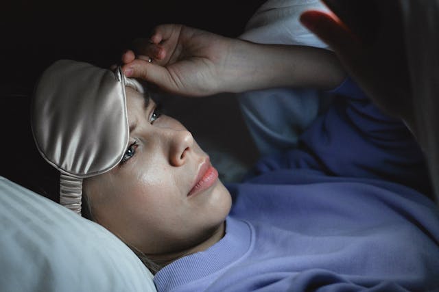 Una mujer en la cama mira por debajo de su antifaz y consulta su teléfono. 