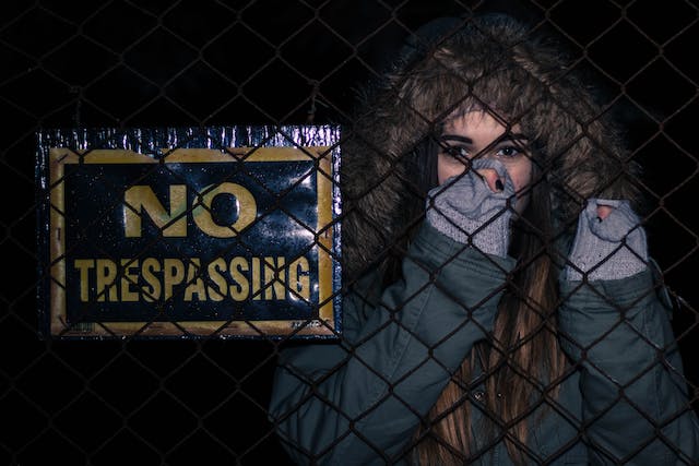 A woman in a parka jacket stands beside a “No Trespassing” sign behind a wire fence. 
